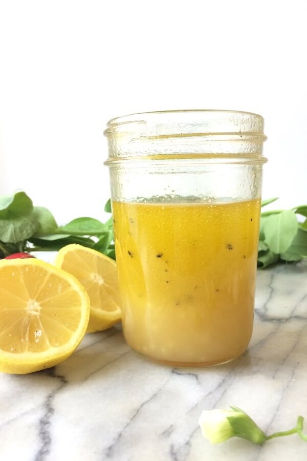 A mason jar filled with lemon-garlic dressing, surrounded by pea shoots and sliced lemons.