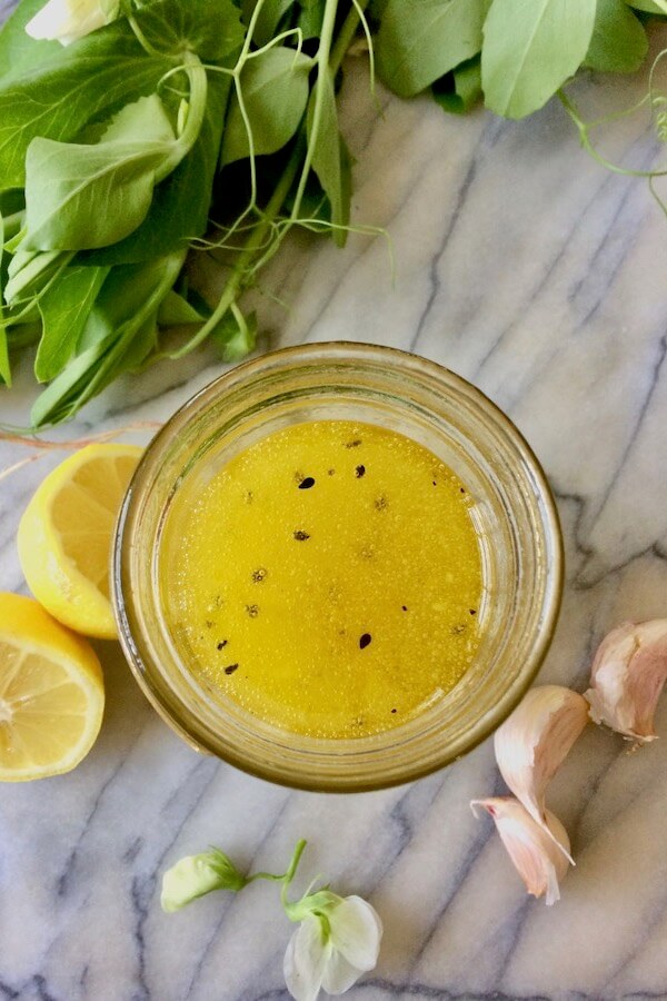 An overhead shot of a jar of lemon-garlic dressing, surrounded by pea shoots, lemons and garlic cloves.